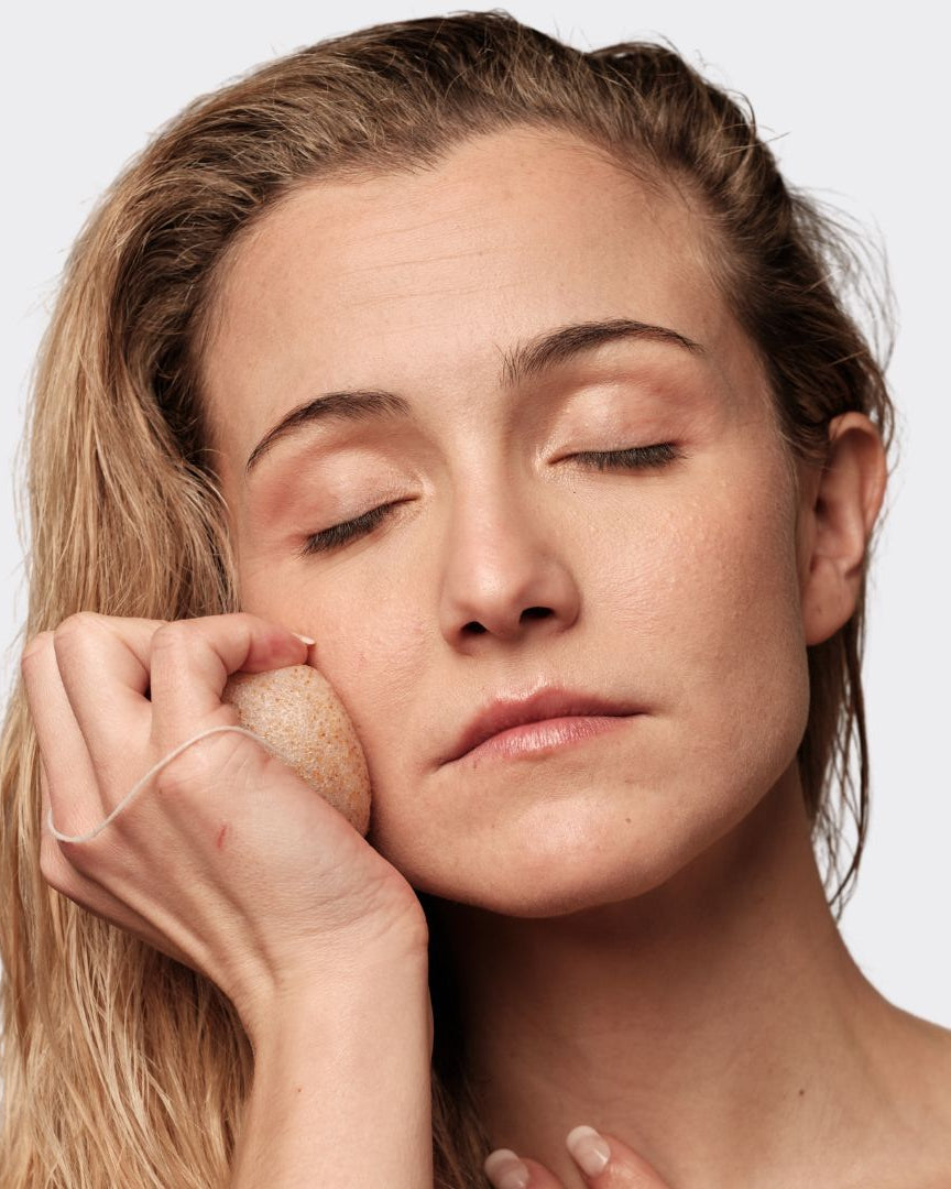 Mujer con ojos cerrados usando una esponja facial Konjac con nuez de Banbu de textura porosa y tono natural.