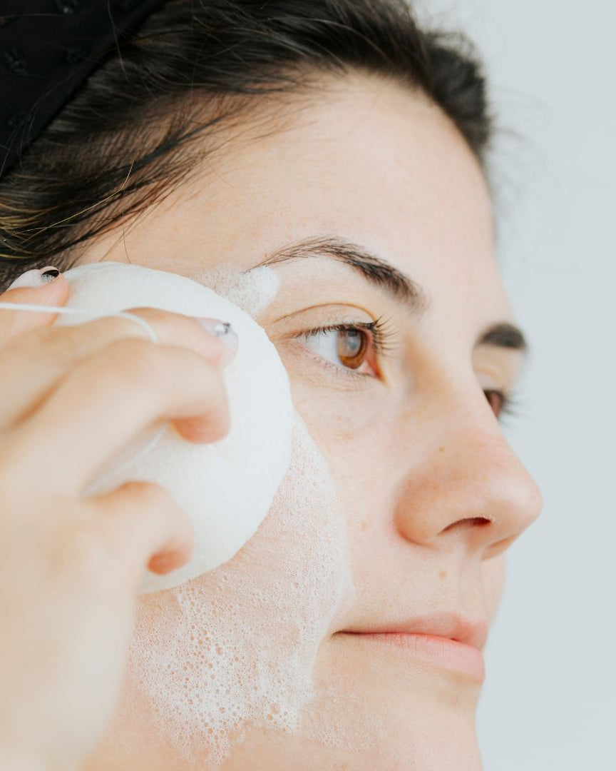 Mujer usando una esponja Konjac blanca y suave de Banbu con espuma para limpiar su rostro con delicadeza.