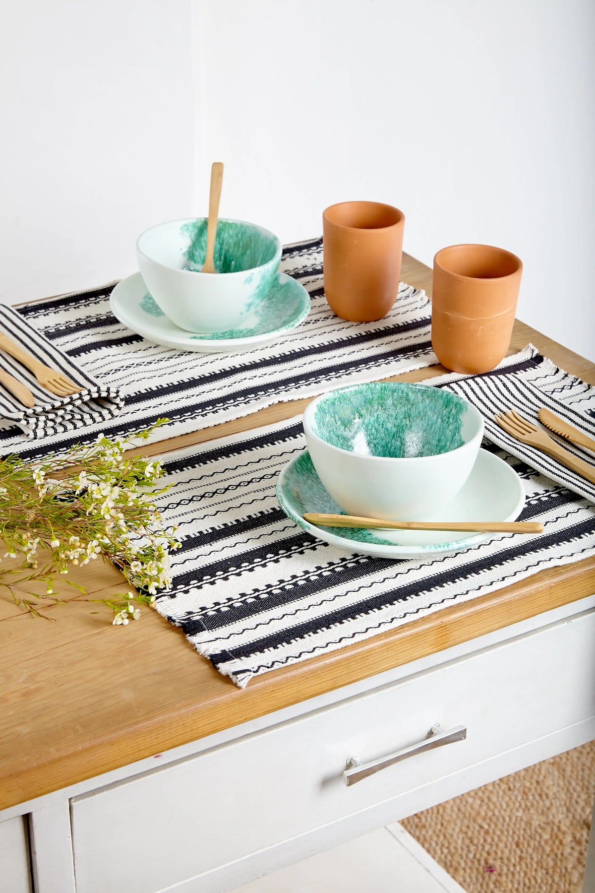Two rectangular black and white patterned woven placemats with fringe, styled on a table with bowls and cups.