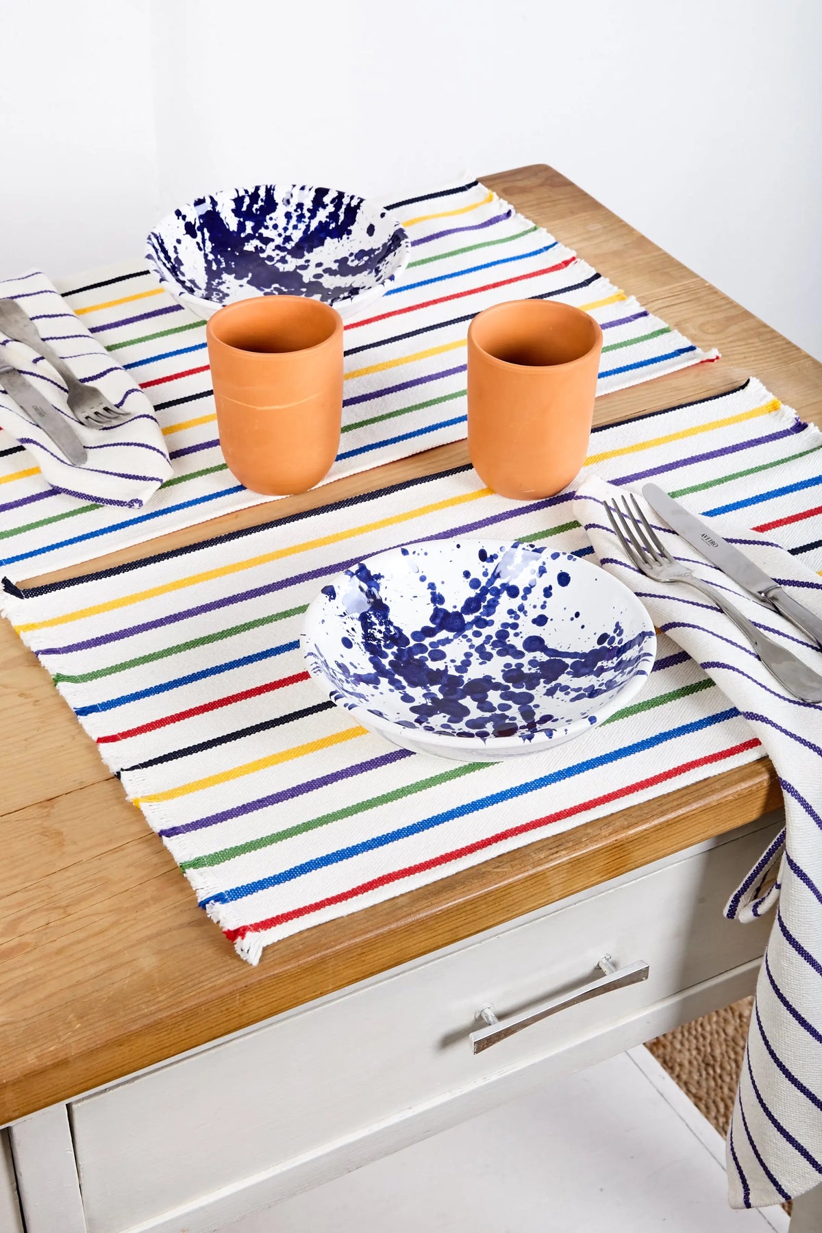 Mantel rayado de colores con platos de cerámica blanca y azul, vasos de terracota y utensilios de acero inoxidable.