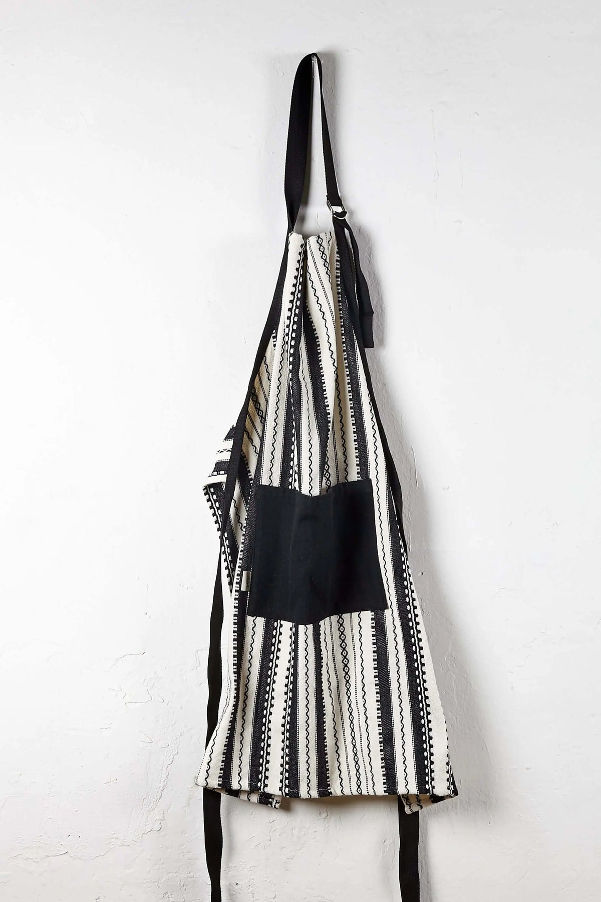Delantal ajustable blanco y negro con estampado de rayas étnicas y bolsillo central liso, colgado sobre pared blanca.