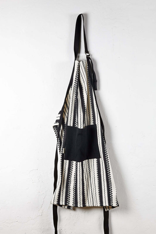 Delantal ajustable blanco y negro con estampado de rayas étnicas y bolsillo central liso, colgado sobre pared blanca.