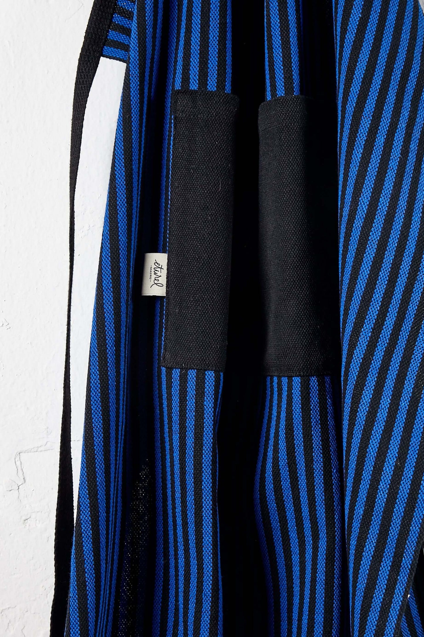Delantal de loneta con patrón de rayas verticales azules y negras y bolsillo frontal grande. Textura resistente.