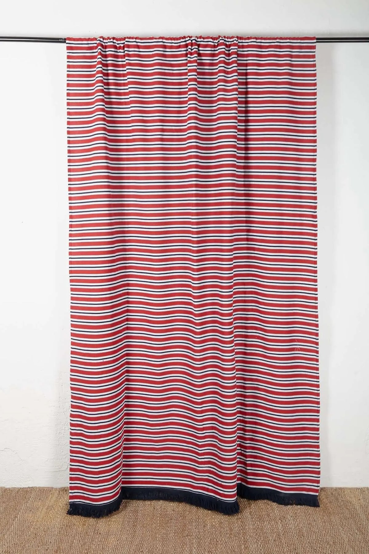 Cortina de loneta con estampado de rayas horizontales rojas, blancas y azul marino, rematada con flecos en el borde.