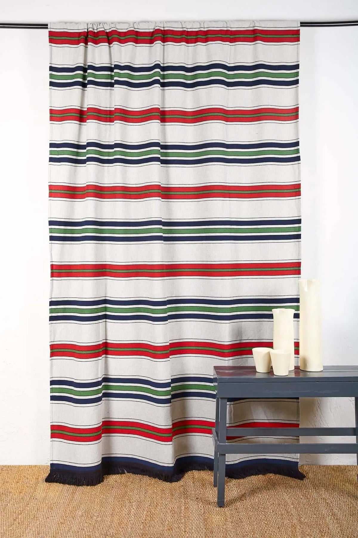 Cortina de tejido artesanal con rayas horizontales en azul, rojo y verde sobre fondo crudo y acabado con flecos.