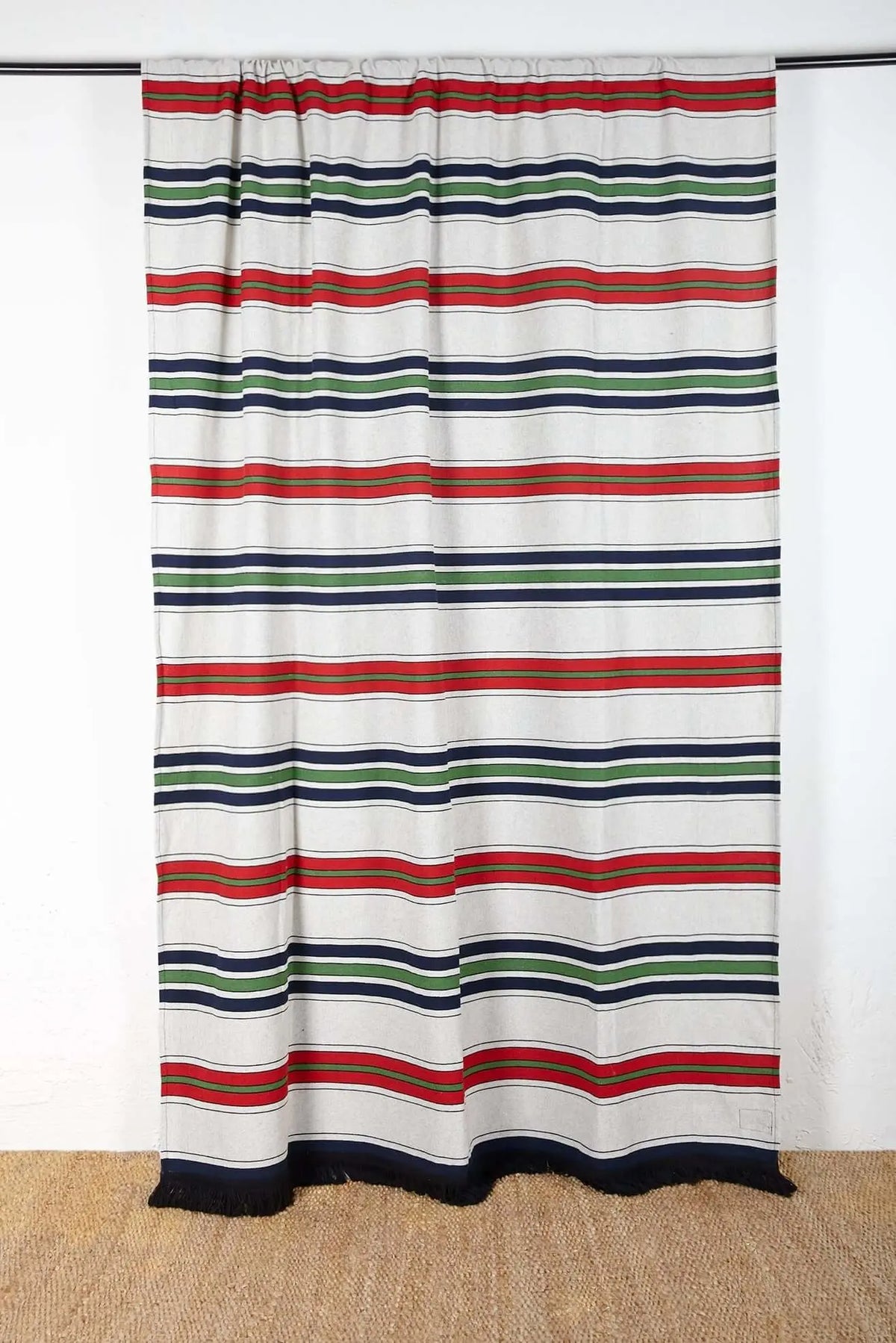 Cortina de tejido canario con rayas horizontales en rojo, verde y azul sobre fondo blanco, acabado con flecos inferiores.