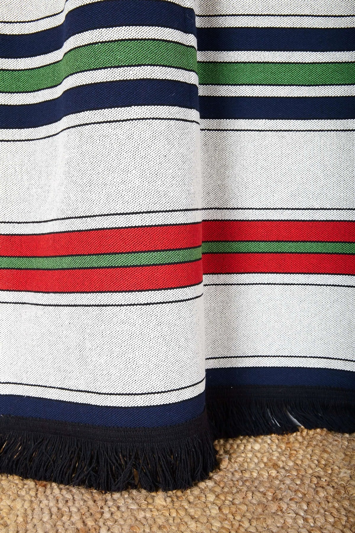 Cortina de tejido canario con rayas horizontales en azul, rojo, verde y blanco, terminada con flecos negros decorativos.
