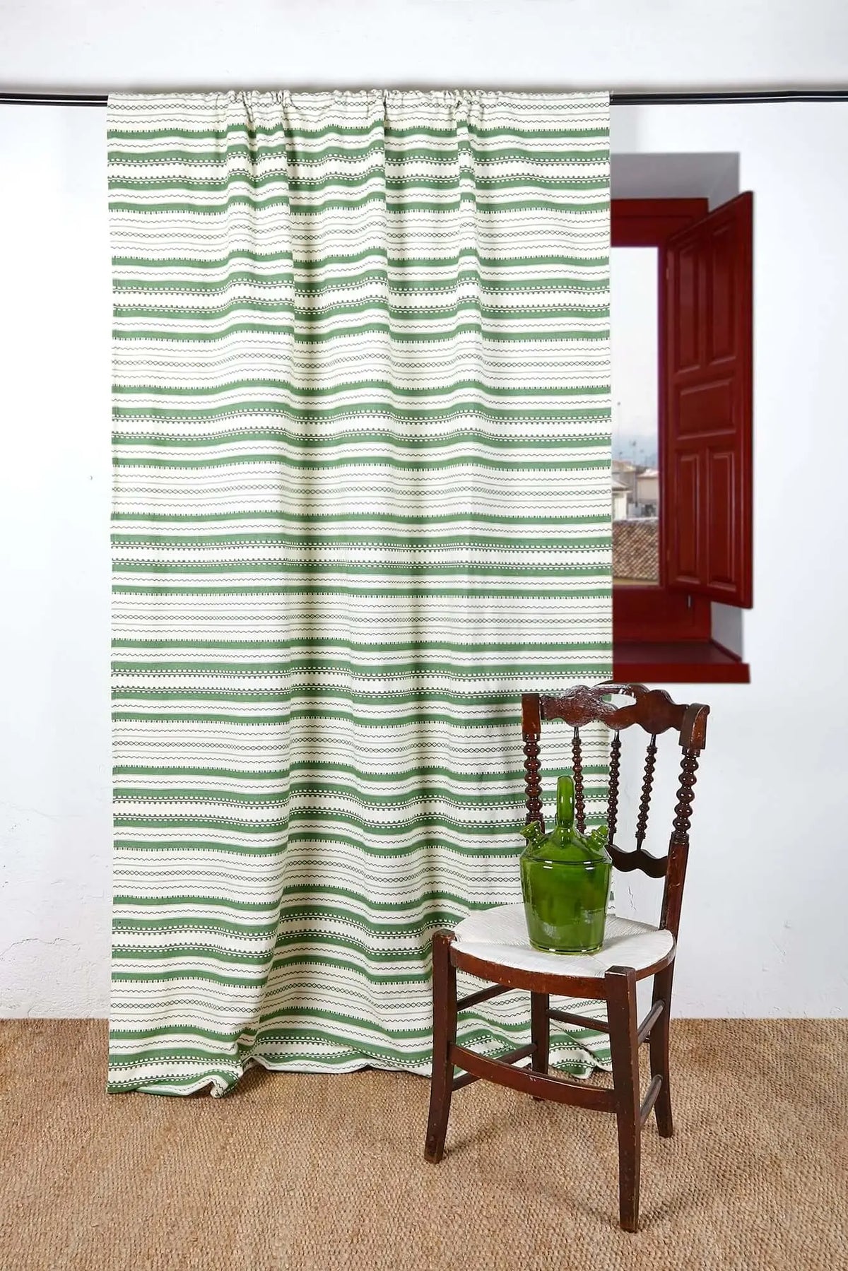 Artisanal green and white striped curtain hanging over a rustic wooden chair in a room with a red window frame.