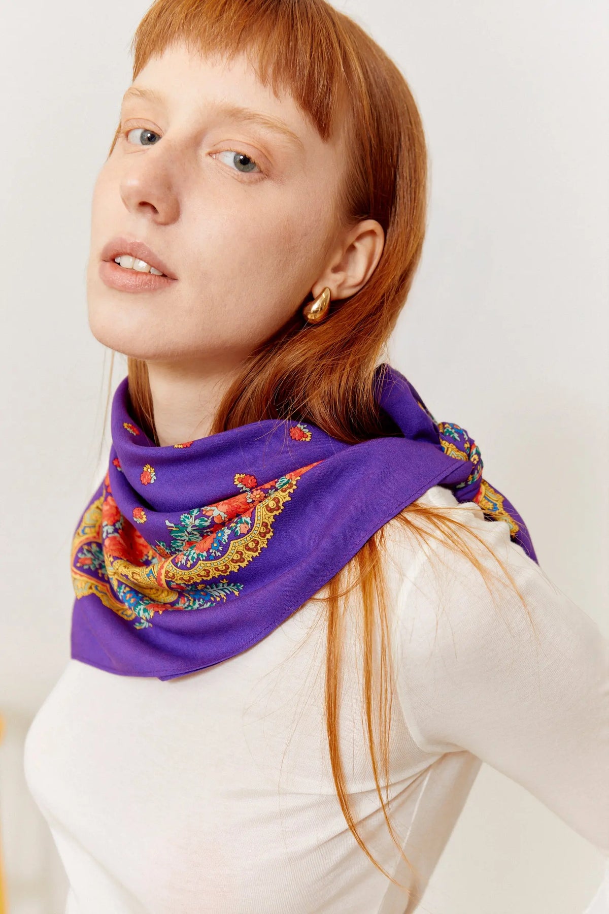 Purple silk scarf with ornate golden chain and floral patterns worn tied around the neck by a redhead woman.