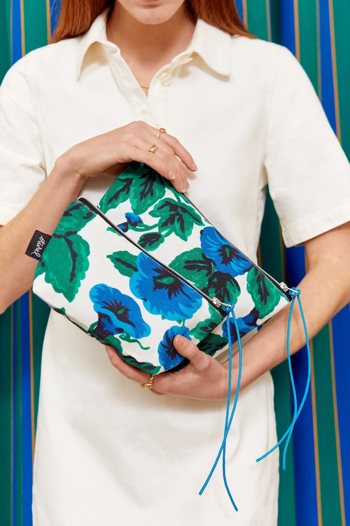 Neceser de lona blanca con estampado de flores azules y hojas verdes, cierre de cremallera y tiradores de cordón azul.