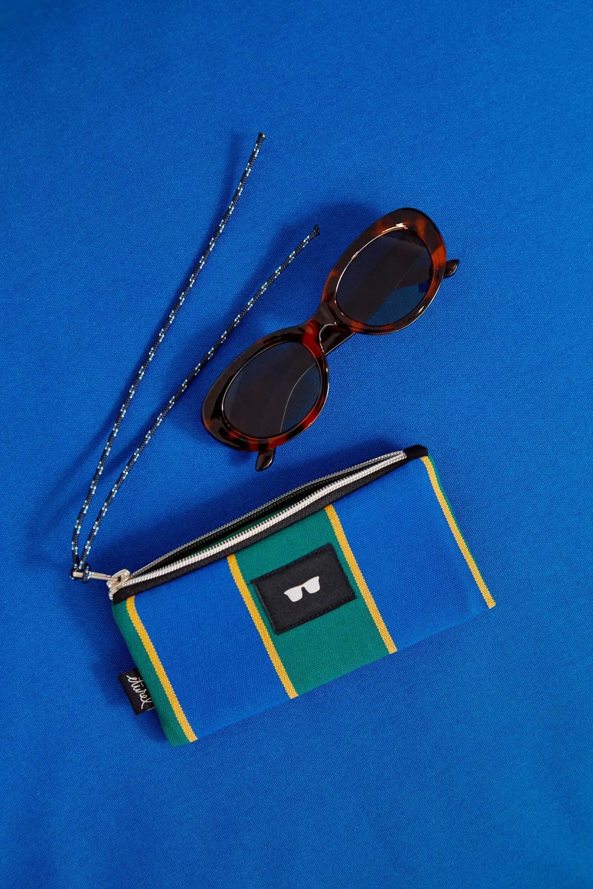 Funda para gafas de lona con rayas azules, verdes y amarillas, junto a unas gafas de sol con cordón sobre fondo azul.