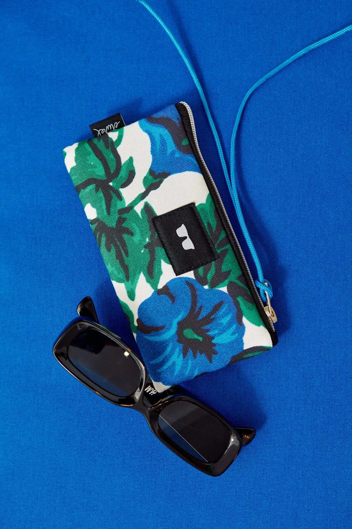 Funda de lona rectangular con estampado floral azul y verde, cremallera, cordón azul y gafas de sol sobre fondo azul.