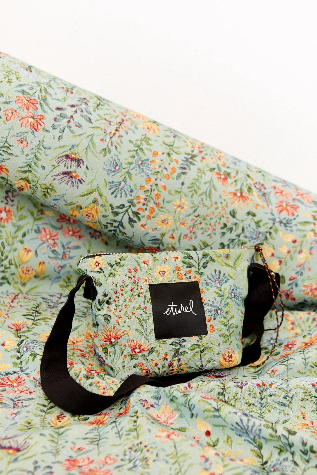 Bolso bandolera de tejido jacquard en tono celeste con estampado de flores coloridas y correa negra ajustable.