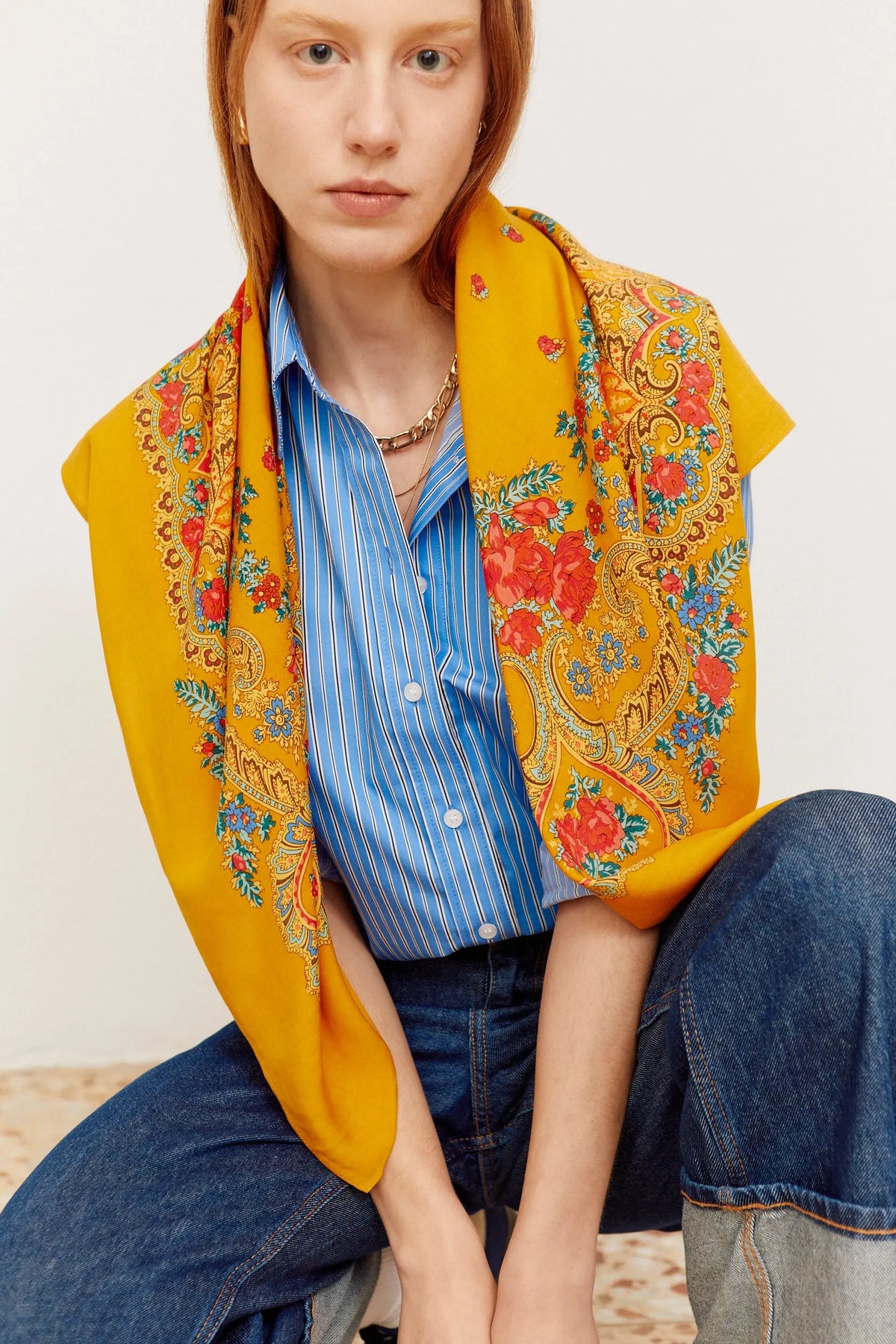 Pañuelo cuadrado ocre con estampado floral y de cadenas, estilo tradicional sobre camisa de rayas azules y jeans.