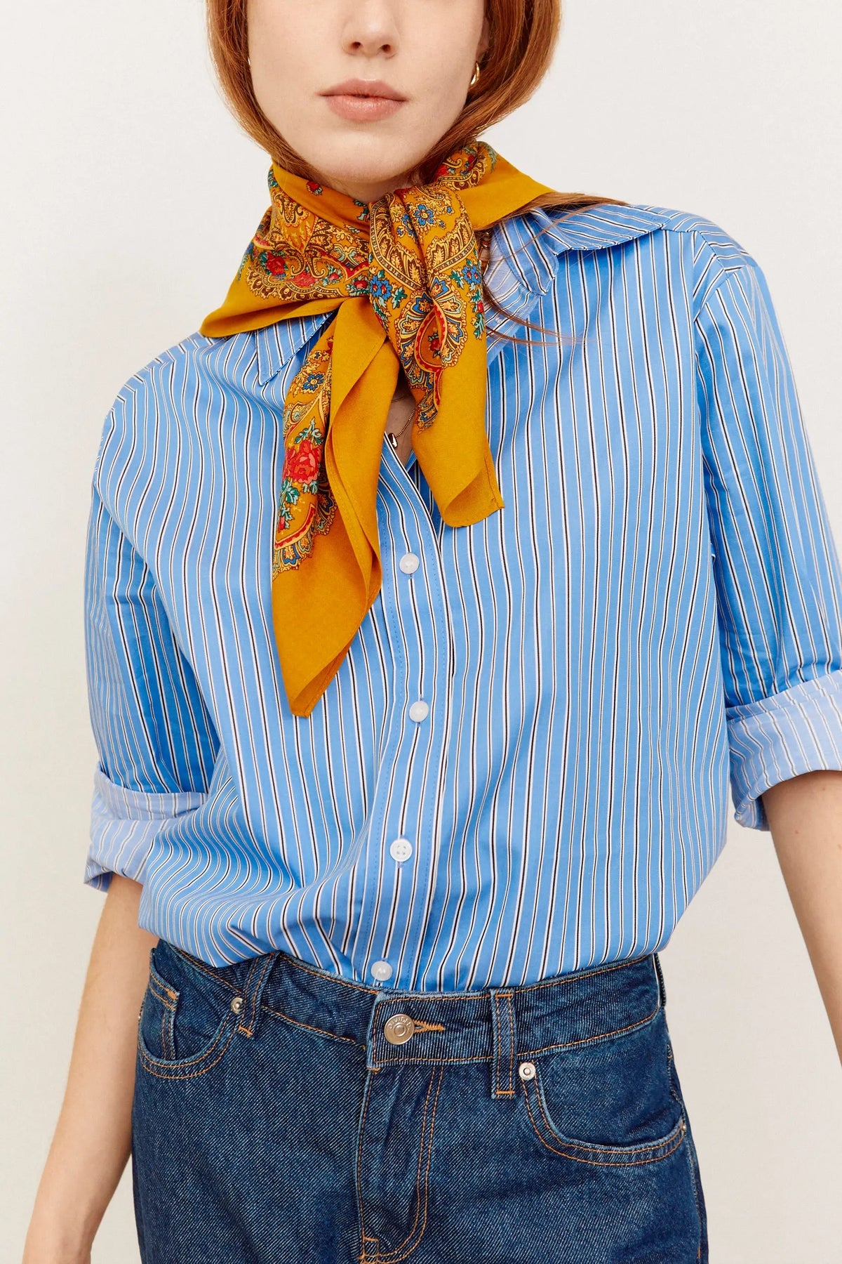 Ochre square scarf with colorful paisley and chain print, tied around neck over a blue striped shirt and jeans.