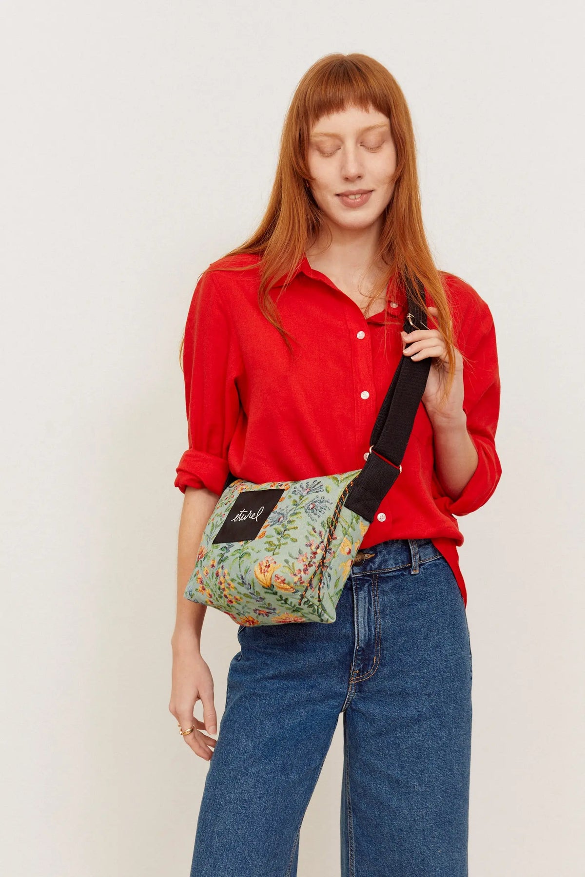 Bolso bandolera de tejido jacquard con estampado floral en tonos celestes y correa negra ajustable, sobre fondo neutro.
