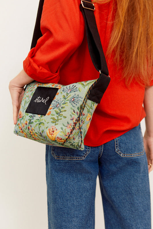 Bolso bandolera de tejido jacquard celeste con estampado de flores, correa negra ajustable y parche decorativo frontal.