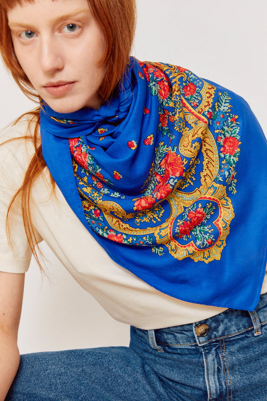 Pañuelo azul con estampado tradicional de flores rojas y motivos en cadena dorada sobre modelo con camiseta blanca.