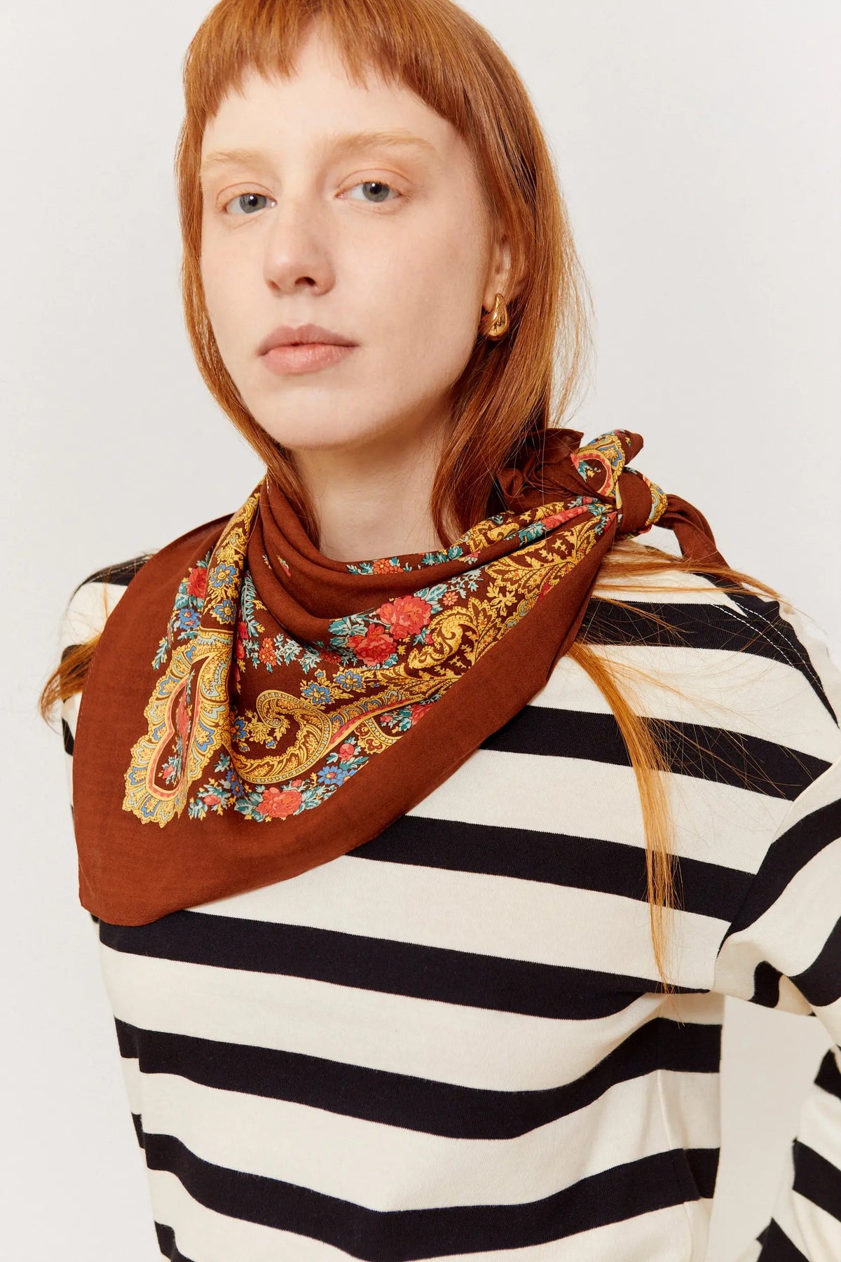 Pañuelo color mocca con estampado floral y cadenas doradas, anudado al cuello sobre camiseta de rayas.