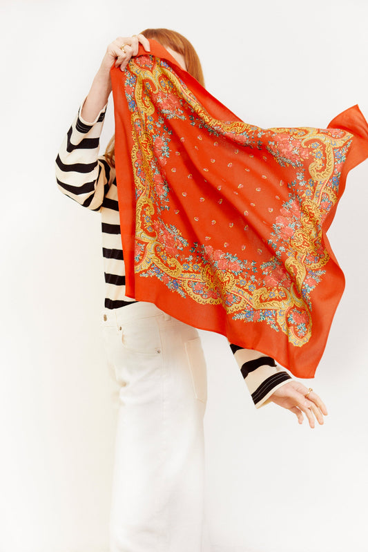 Pañuelo rojo cuadrado con estampado floral y de cadenas doradas, estilo vintage, sostenido por una mujer de perfil.