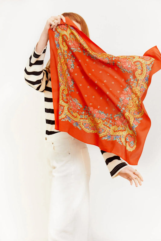 Pañuelo rojo cuadrado con estampado floral y de cadenas doradas, estilo vintage, sostenido por una mujer de perfil.