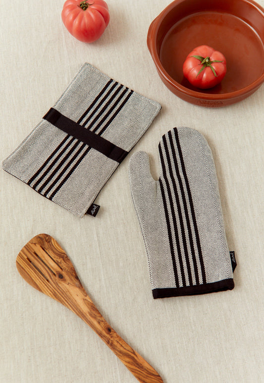 Guantes de cocina gris y negro con rayas, de algodón, junto a un utensilio de madera y tomates rojos.