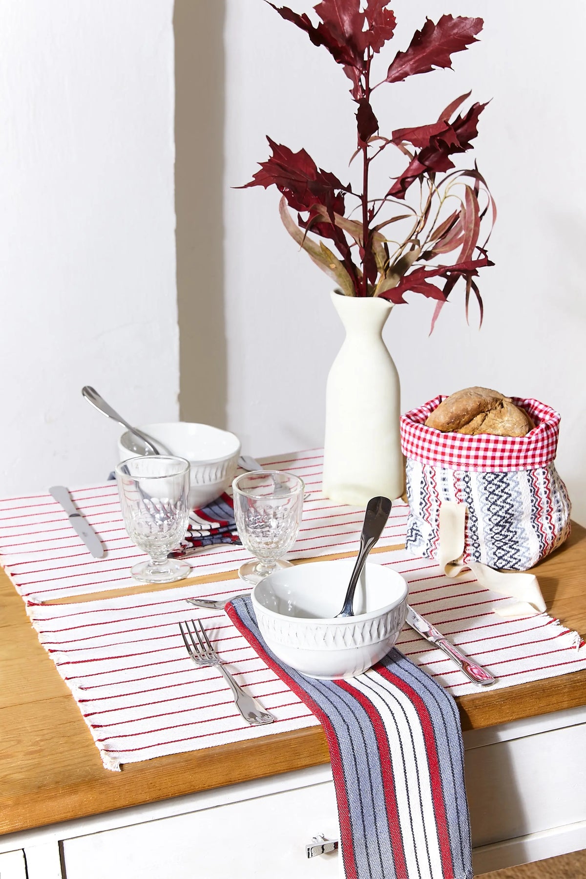 Pack de 2 manteles individuales de loneta blanca con rayas granates sobre una mesa de madera con vajilla y decoración.