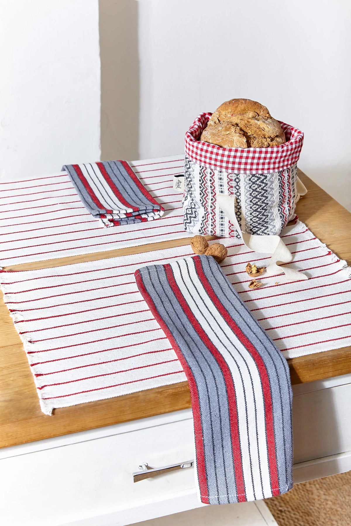 Two white canvas placemats with thin red stripes, featuring folded, multi-colored striped napkins and a bread basket.