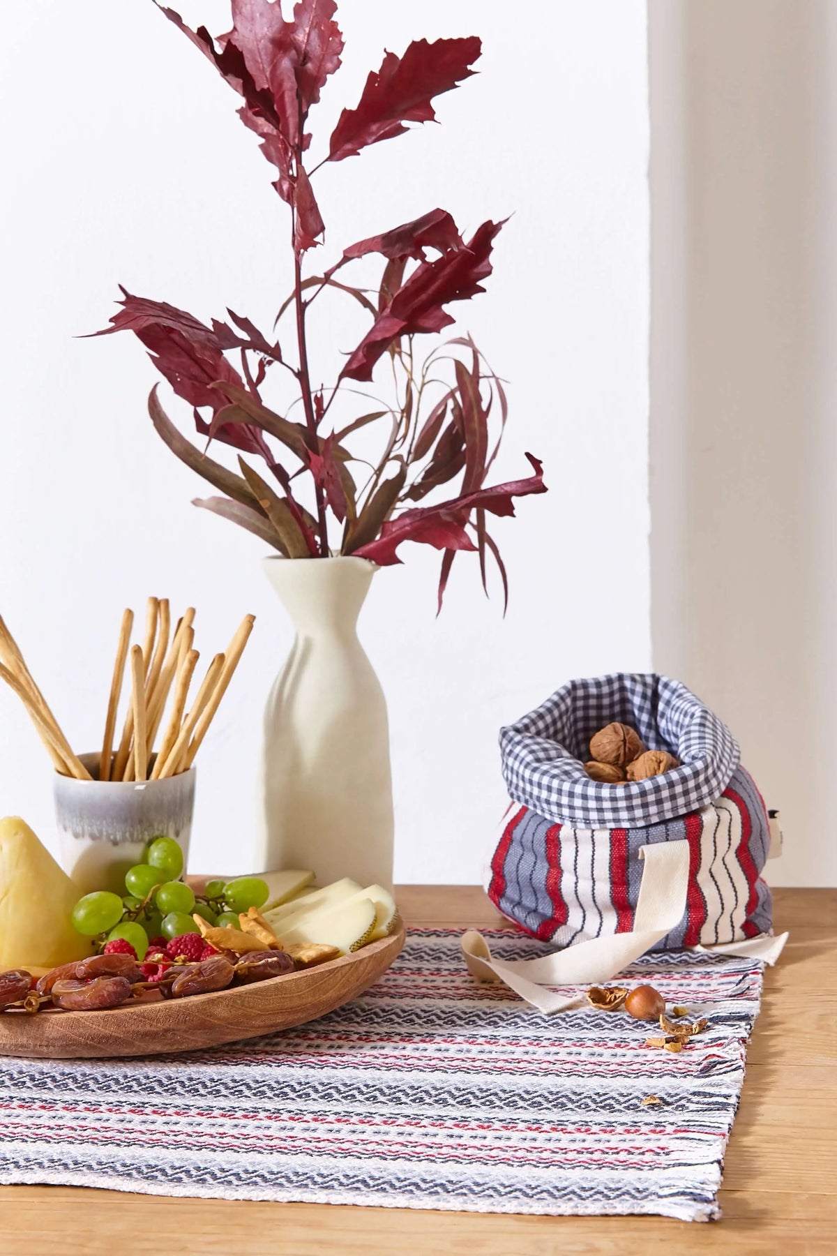 Camino de mesa artesanal granate con tejido a rayas sobre madera, junto a bandeja de aperitivos y decoración otoñal.
