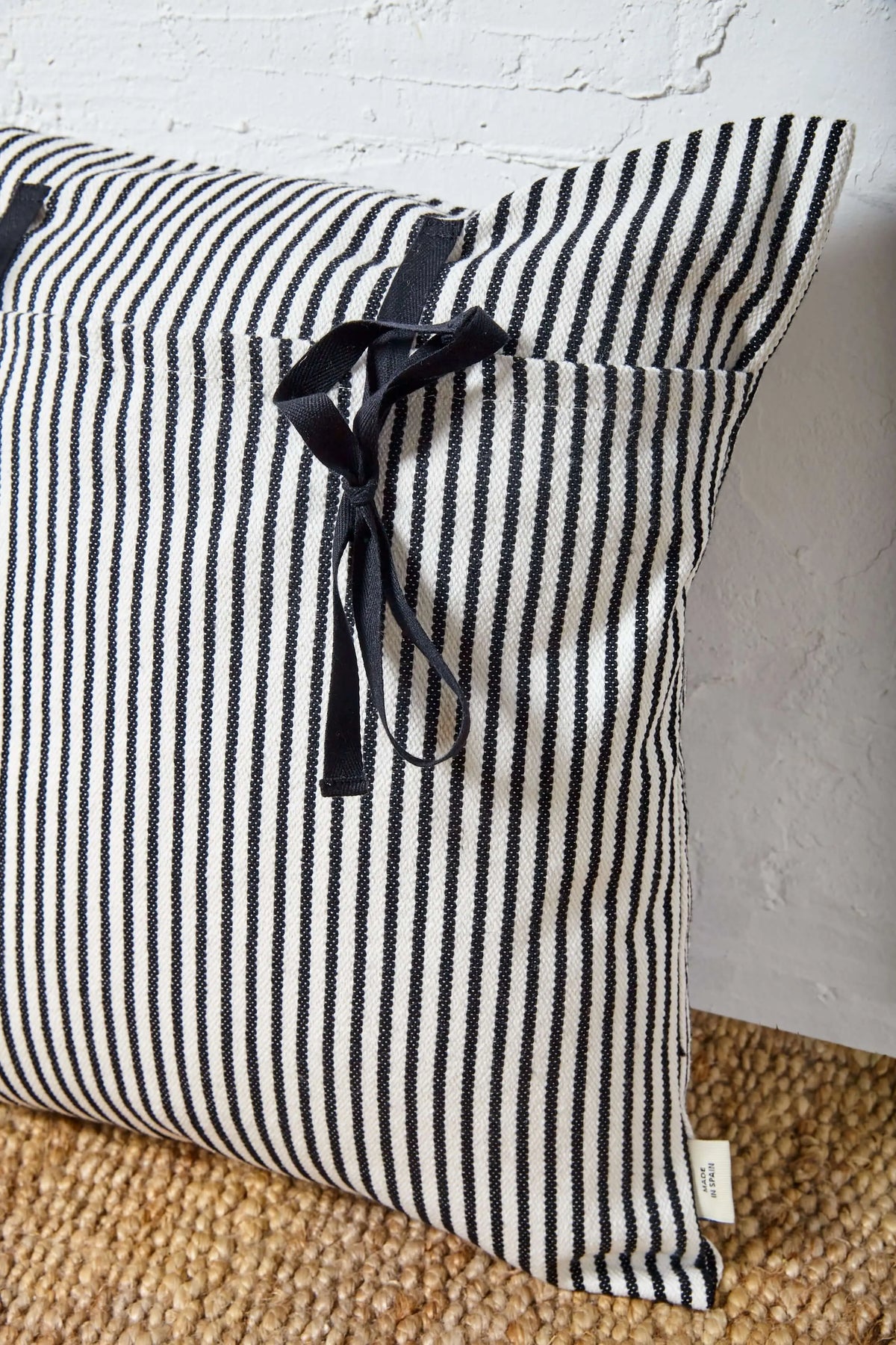 Funda de cojín cuadrada de loneta con rayas verticales blanco y negro, cierre con lazos negros y textura rústica.