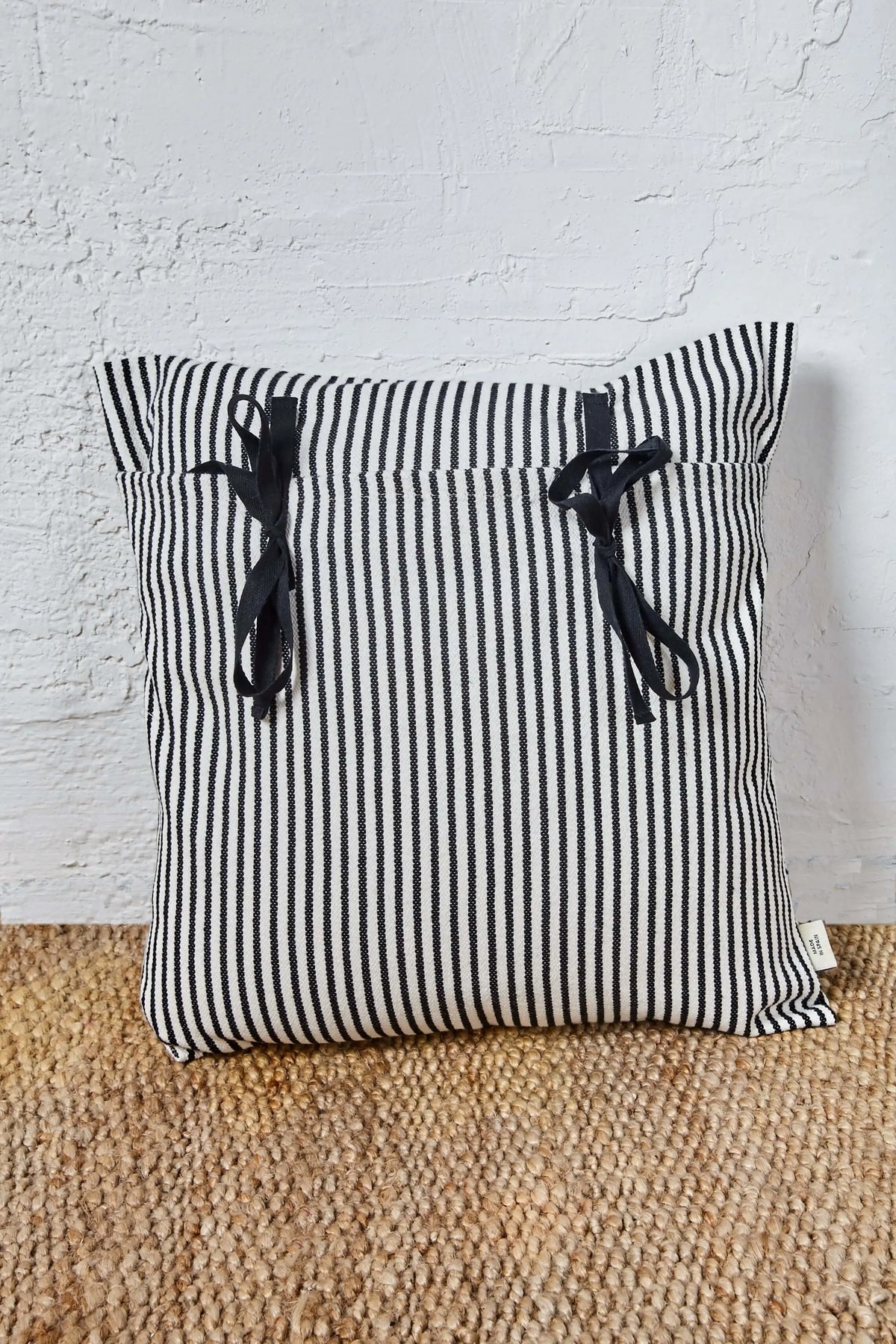 Funda de cojín de loneta a rayas blancas y negras, cuadrada, con dos lazos decorativos negros y cierre tipo estribera.