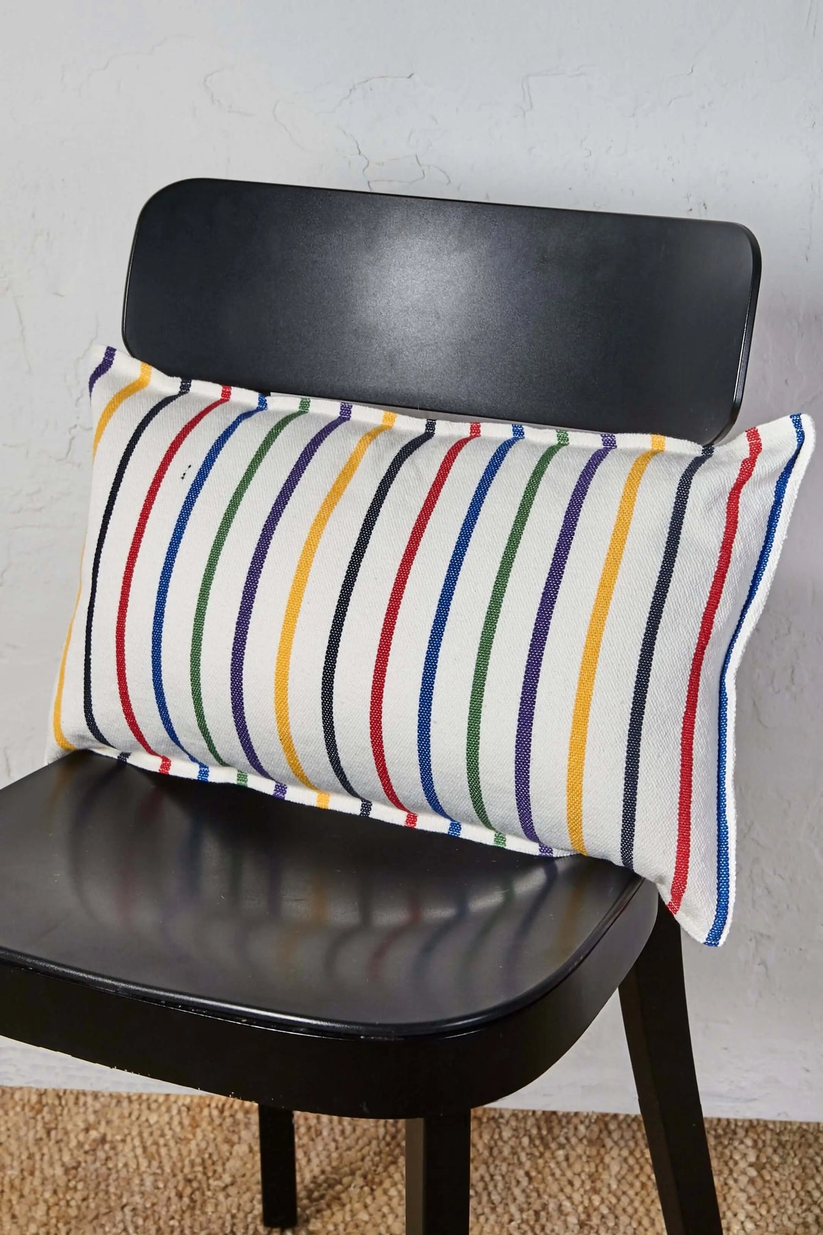Funda de cojín rectangular de 30x50 cm, tejido blanco con rayas verticales de colores sobre una silla negra.