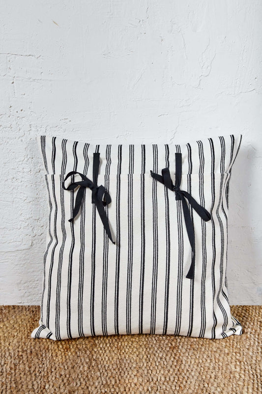 Funda de cojín de loneta a rayas blancas y negras, cuadrada, con dos lazadas decorativas de tela negra en el frente.