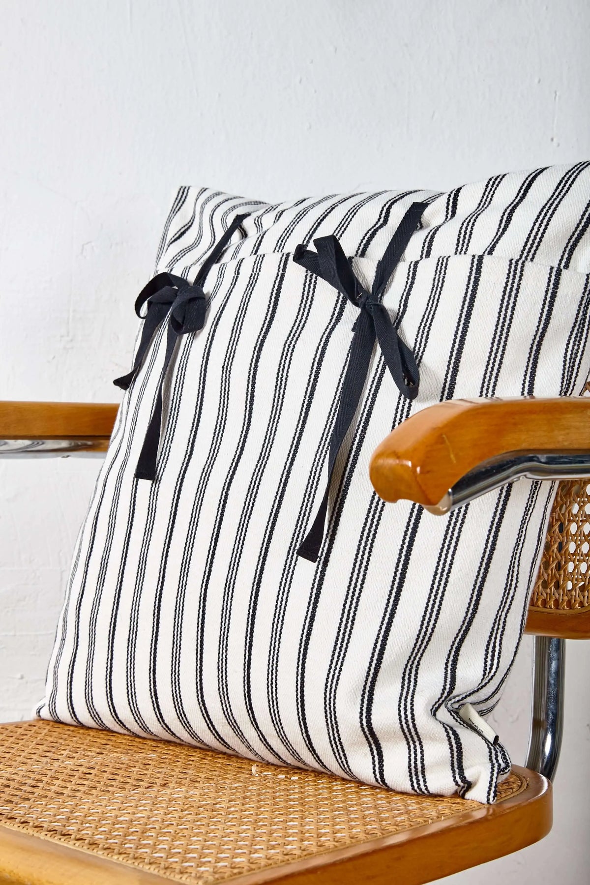 Funda de cojín de loneta blanca con rayas negras verticales y dos lazos decorativos negros, de 50x50 cm, sobre una silla.