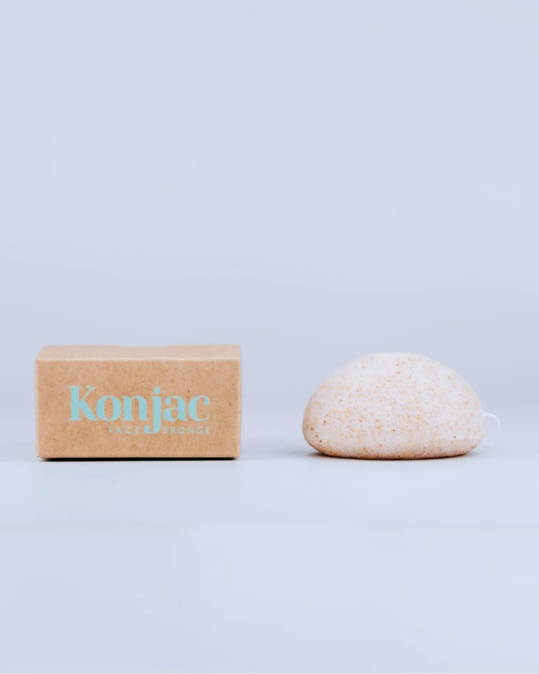 Esponja facial Konjac de Banbu con partículas de nuez junto a su caja de cartón reciclado sobre fondo gris claro.