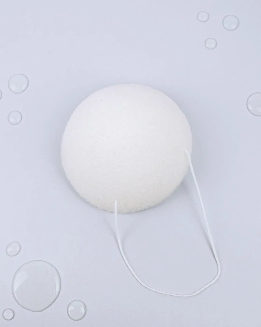 Esponja facial Banbu blanca, redonda y porosa, con cordón para colgar sobre un fondo gris con gotas de agua.