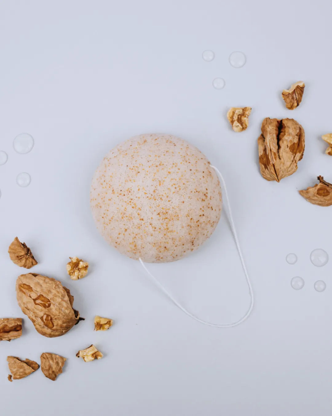 Esponja facial Konjac Banbu con partículas de nuez, textura porosa y color crema sobre fondo gris con trozos de nuez.