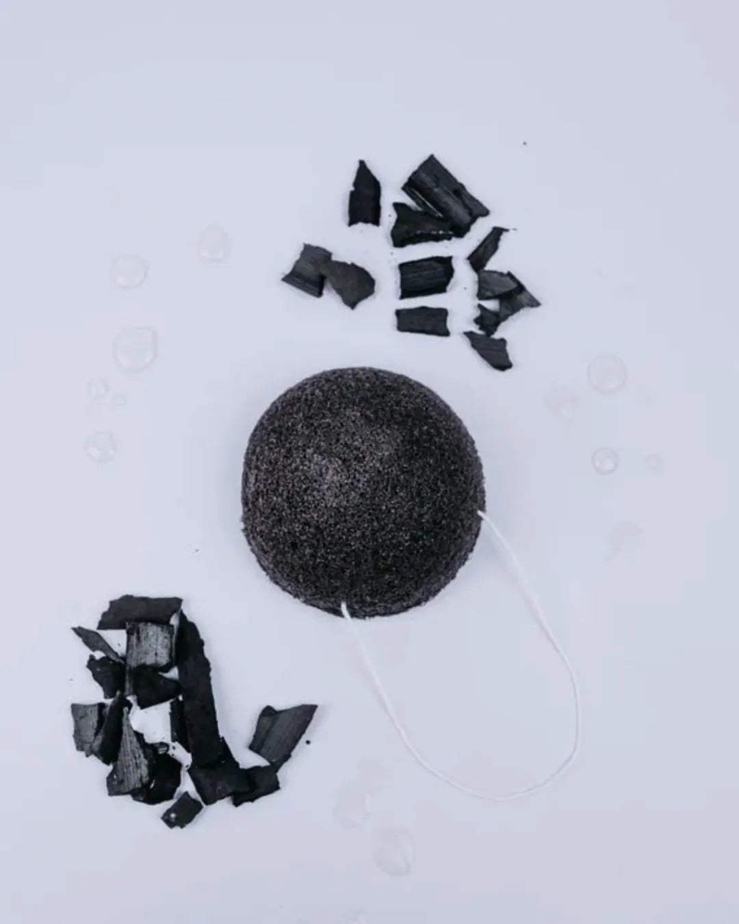 Esponja Konjac negra de Banbu con carbón, rodeada de trozos de carbón y gotas de agua sobre fondo blanco.