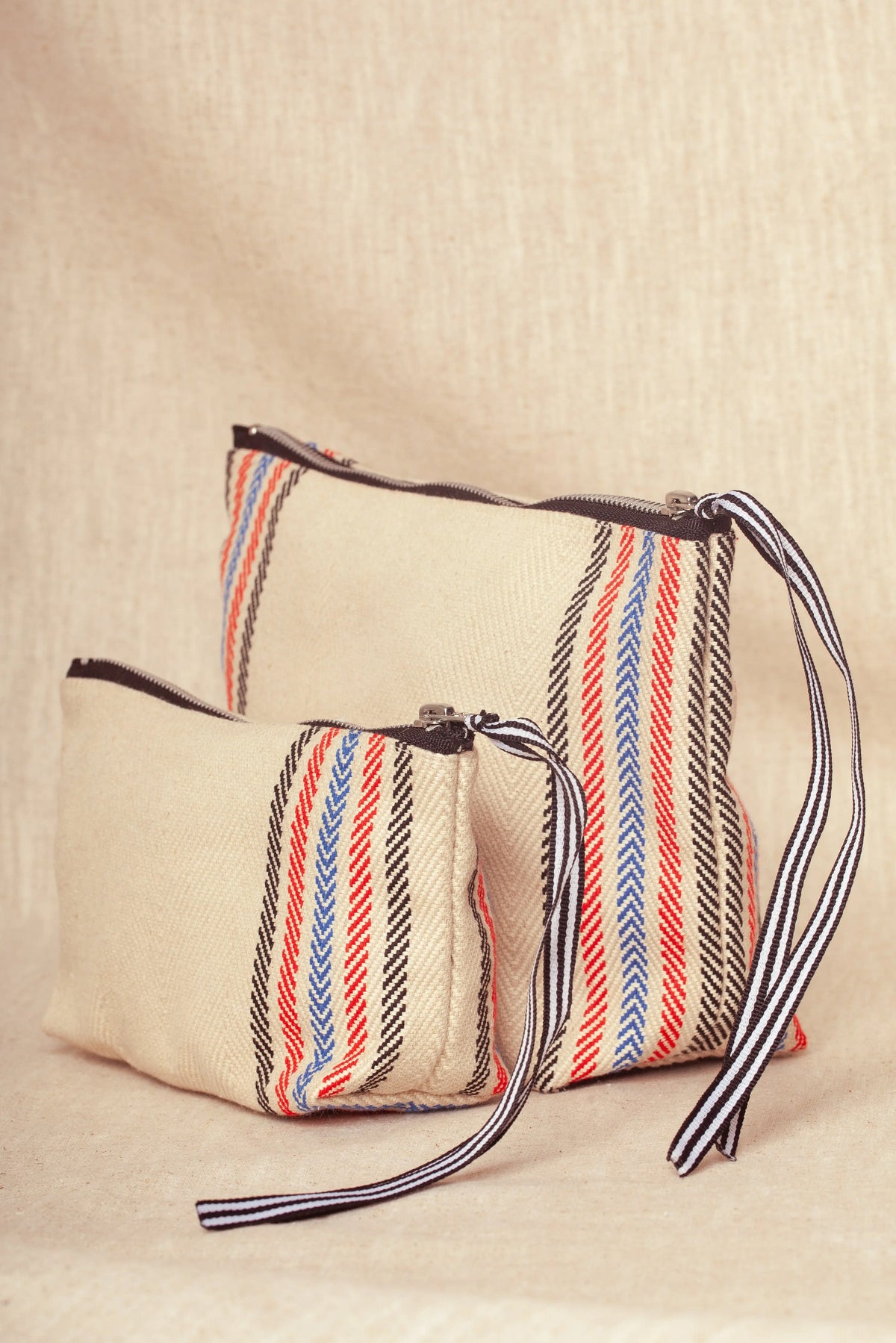Two off-white twill toiletry bags with black, red, and blue striped patterns, black zippers, and striped pull cords.