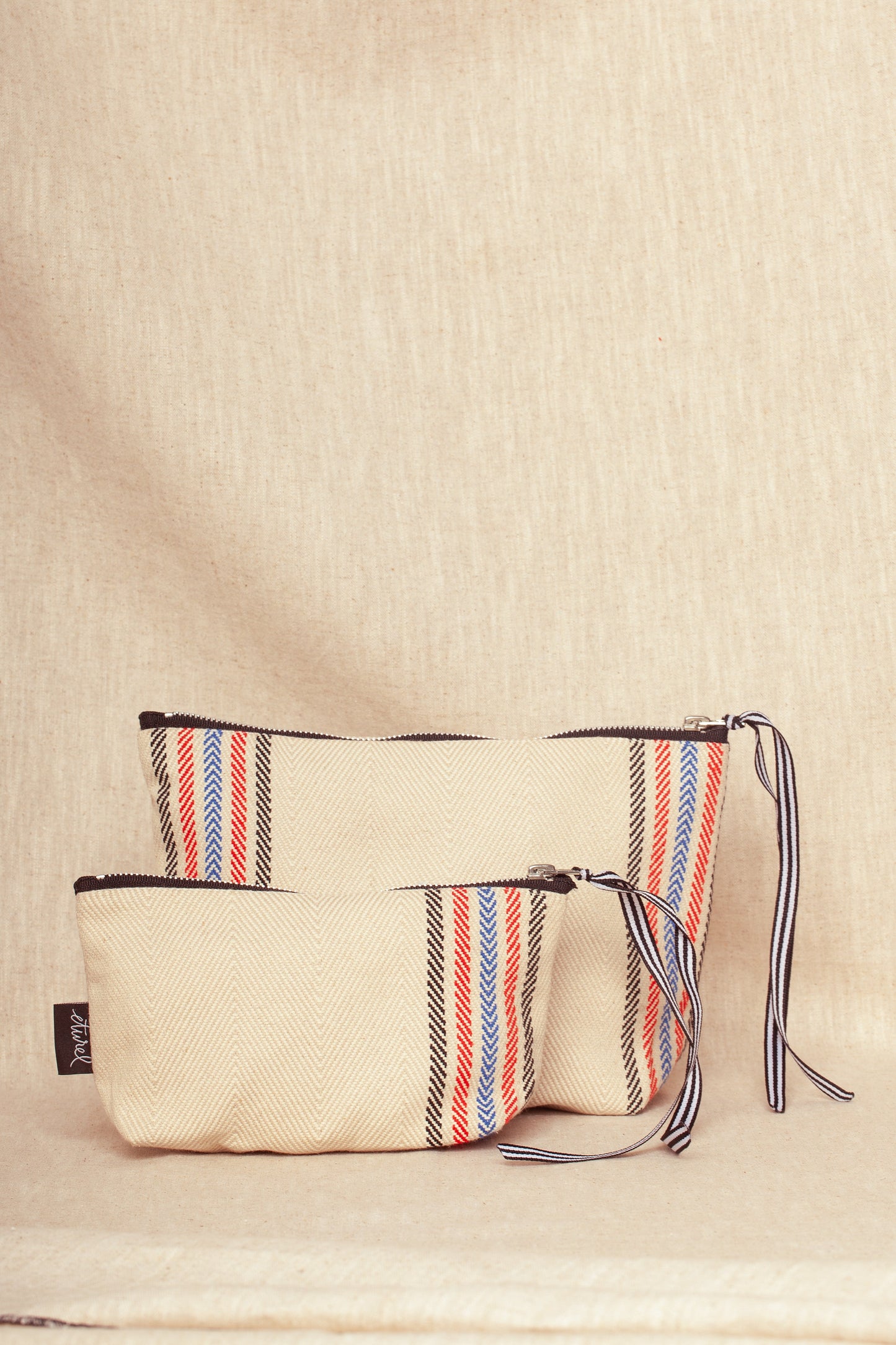 Two cream-colored herringbone weave pouches with red and blue striped accents, black zippers, and ribbon pull tabs.