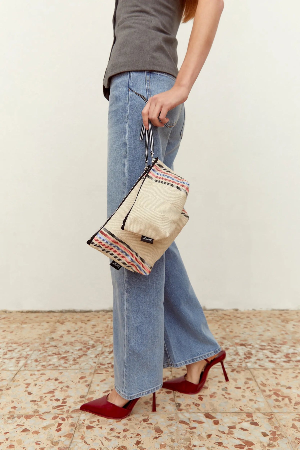 Neceser de sarga color crudo con rayas decorativas, cierre de cremallera y asa corta, sostenido por una mujer.