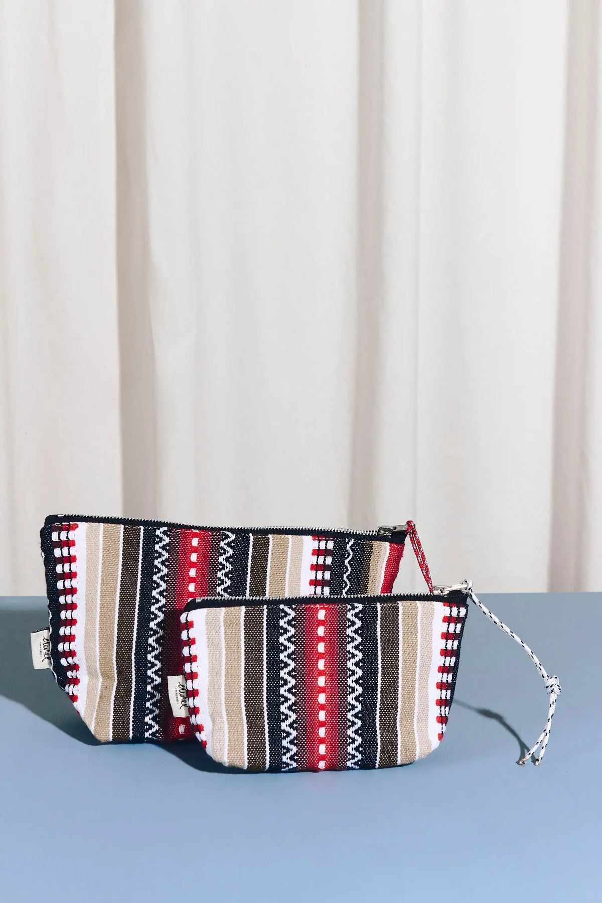 Two zippered pouches with red, black, beige, and white striped woven fabric patterns on a blue surface.