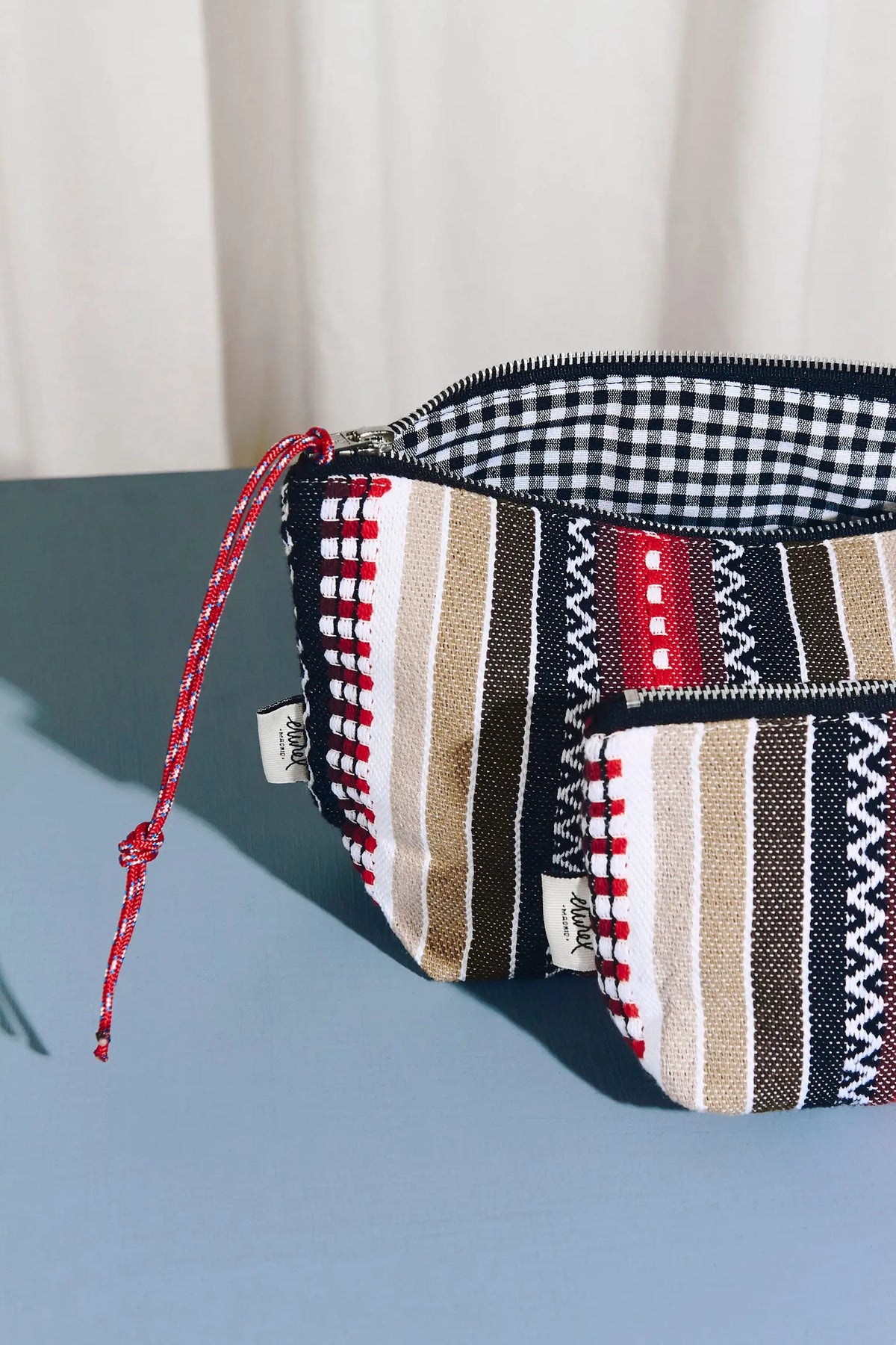 Neceser y estuche con tejido manchego a rayas rojas, negras y beige con interior de cuadros vichy y cierre de cremallera.