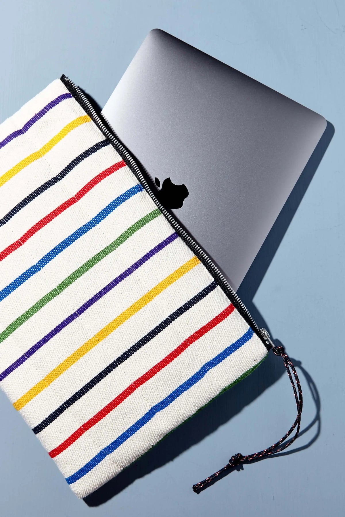 Funda de tela blanca con rayas horizontales multicolores y cremallera negra, conteniendo un ordenador portátil gris.