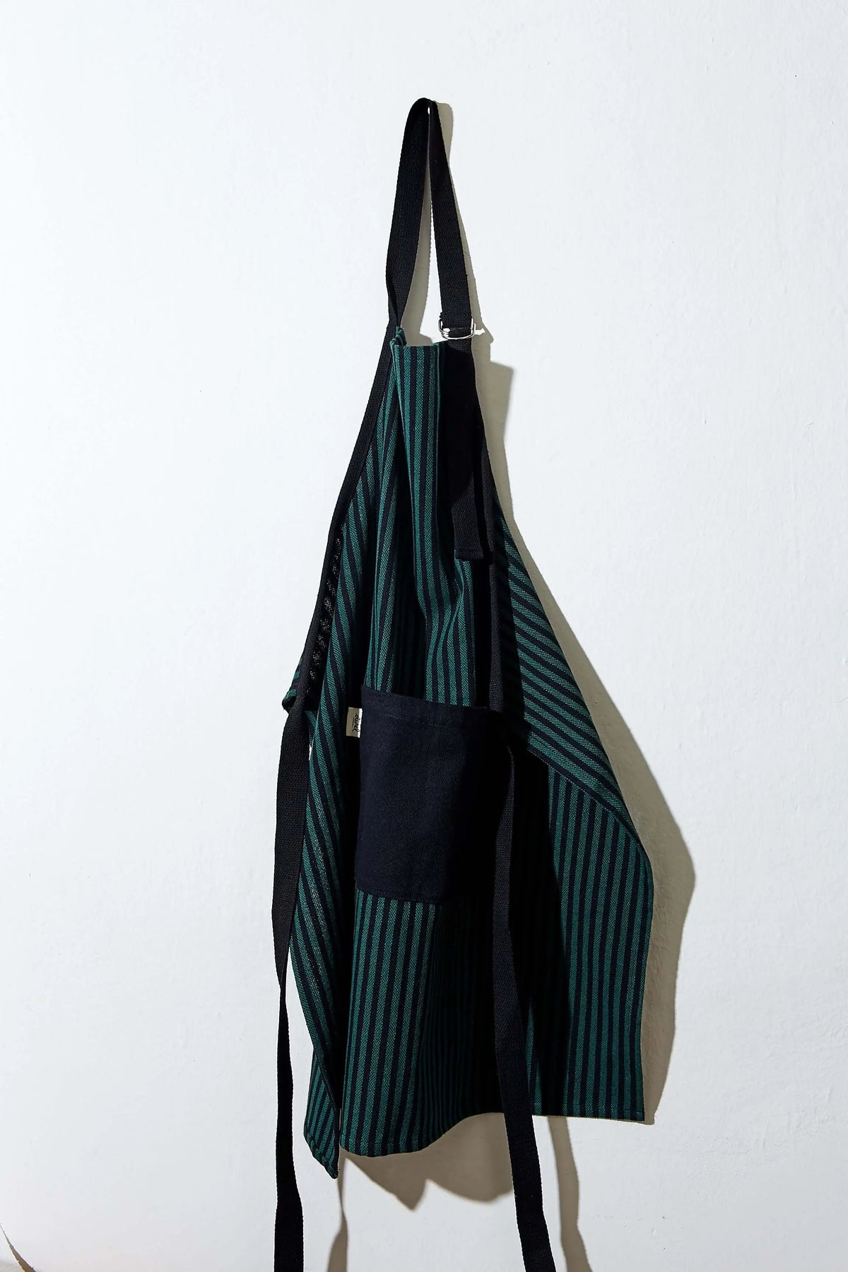 Adjustable green and black striped canvas apron with black ties and a solid black front pocket hanging on a wall.