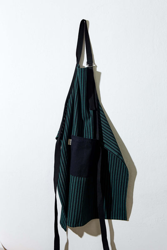 Adjustable green and black striped canvas apron with black ties and a solid black front pocket hanging on a wall.