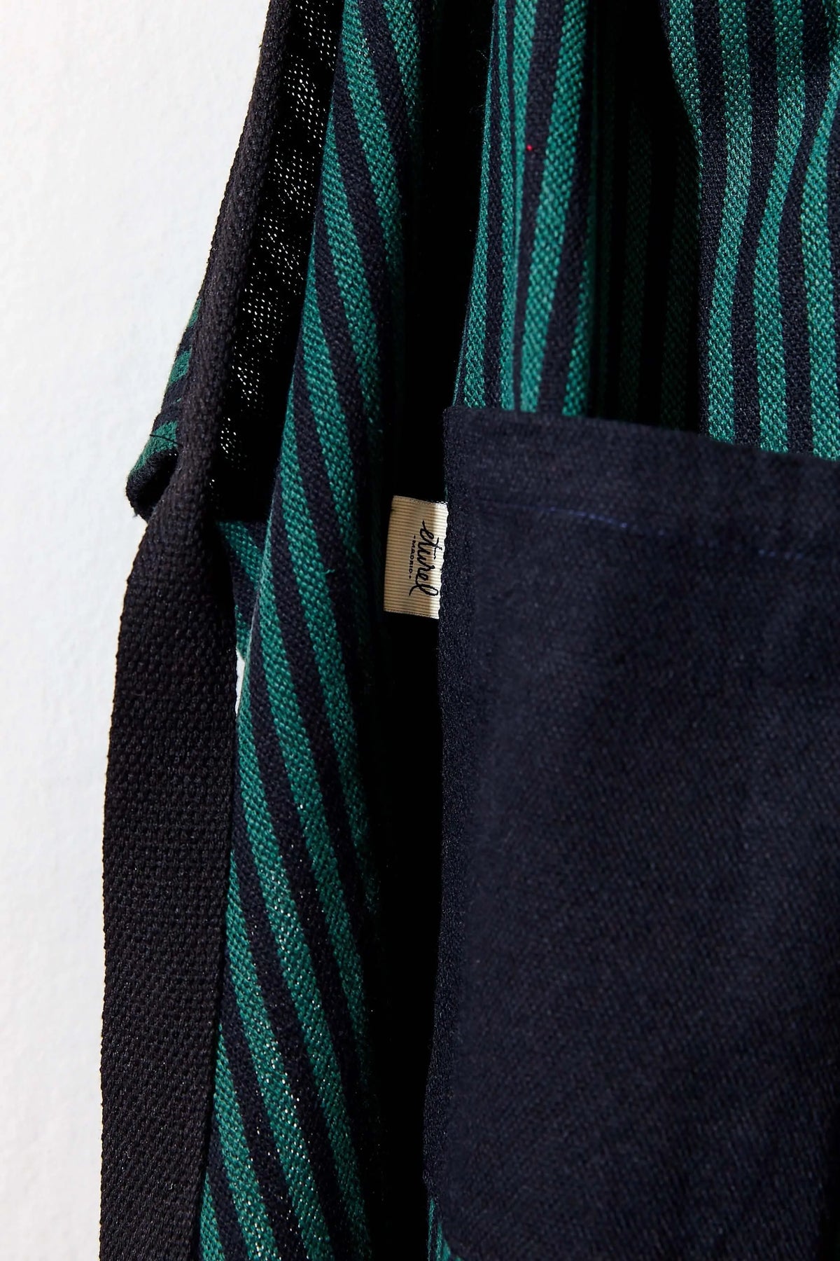 Delantal de loneta a rayas verde y negro, estilo estribera, con bolsillo frontal grande y cinta ajustable negra.