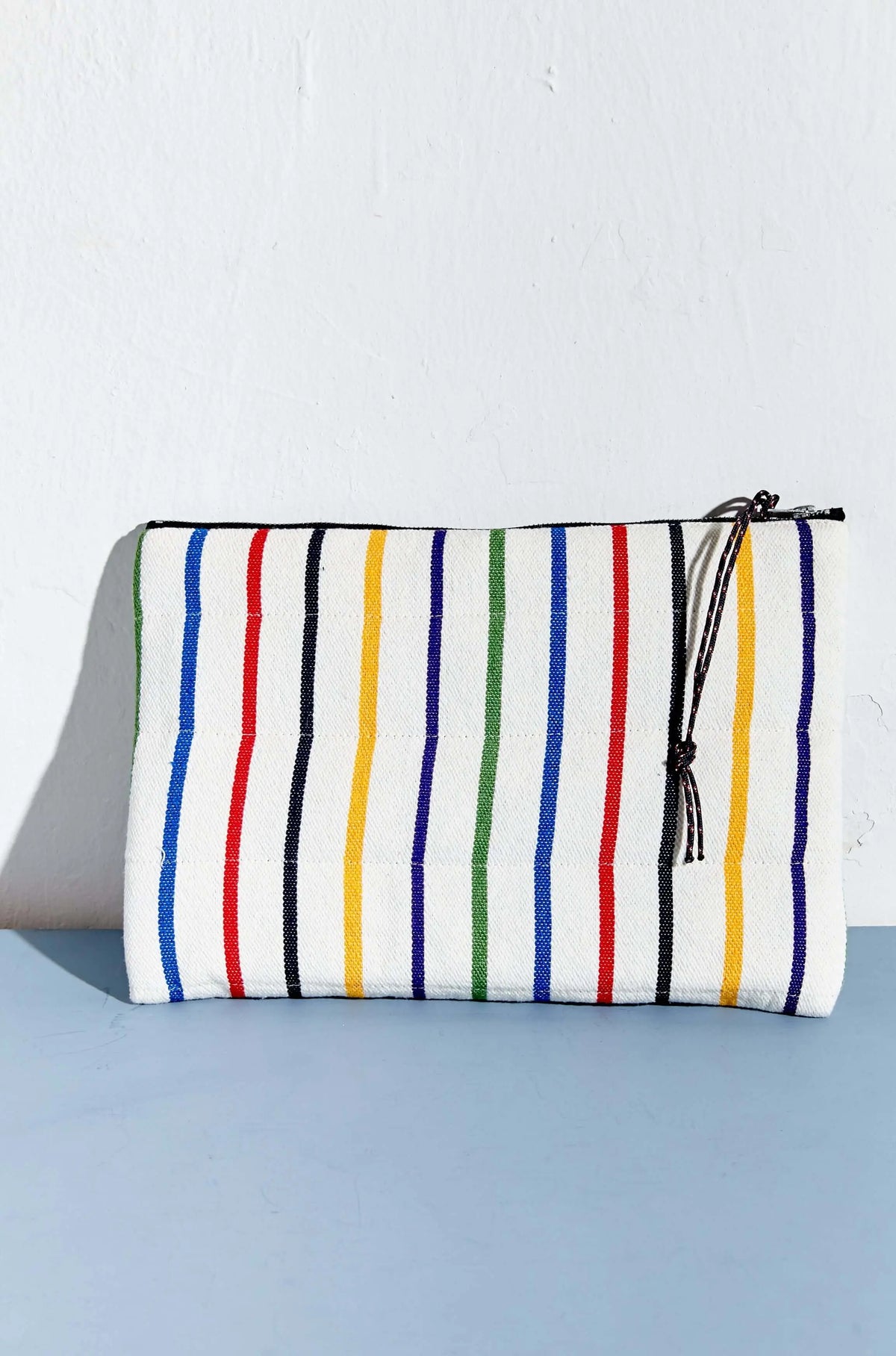 Funda rectangular de tela blanca con rayas verticales de colores, cierre de cremallera y cordón, sobre fondo azul.