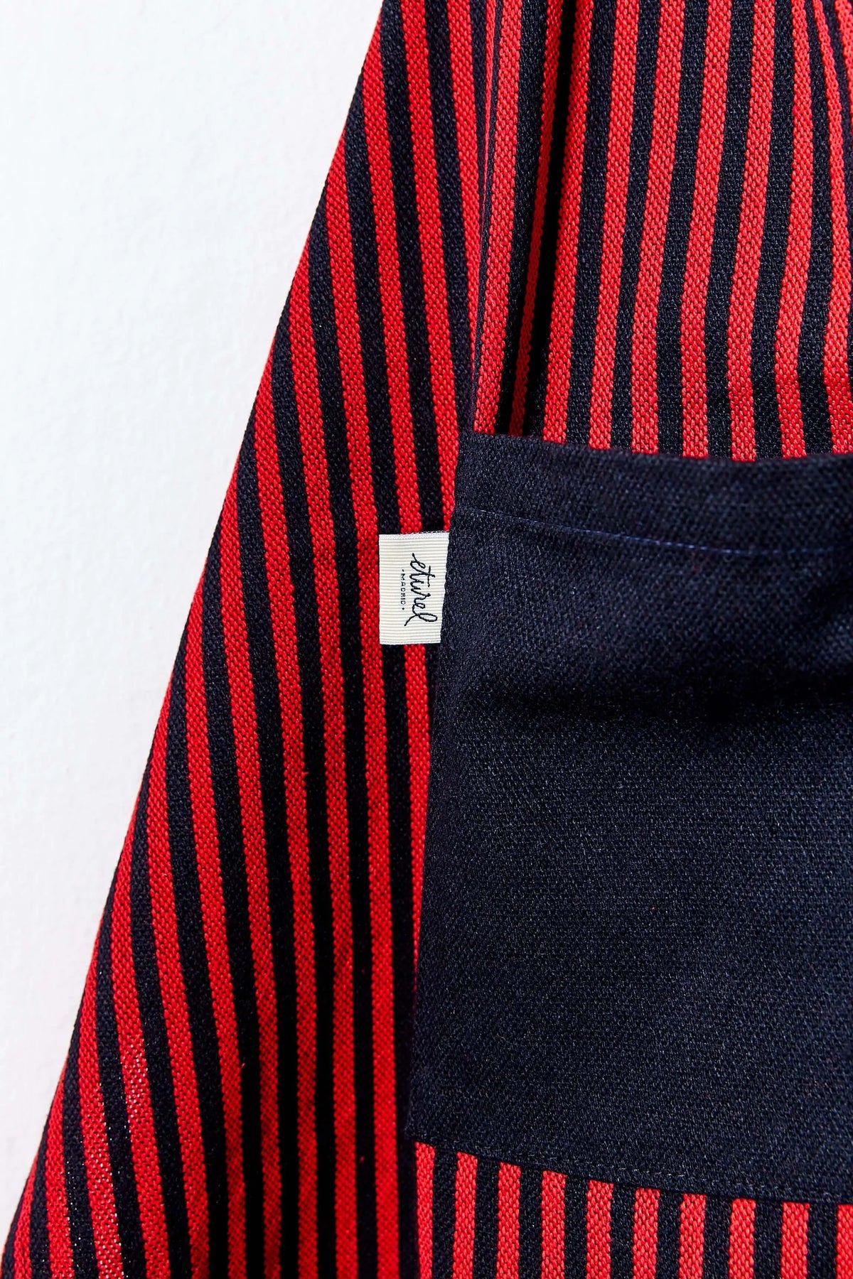 Delantal de loneta con rayas verticales rojas y negras, tejido resistente y bolsillo lateral de color sólido.