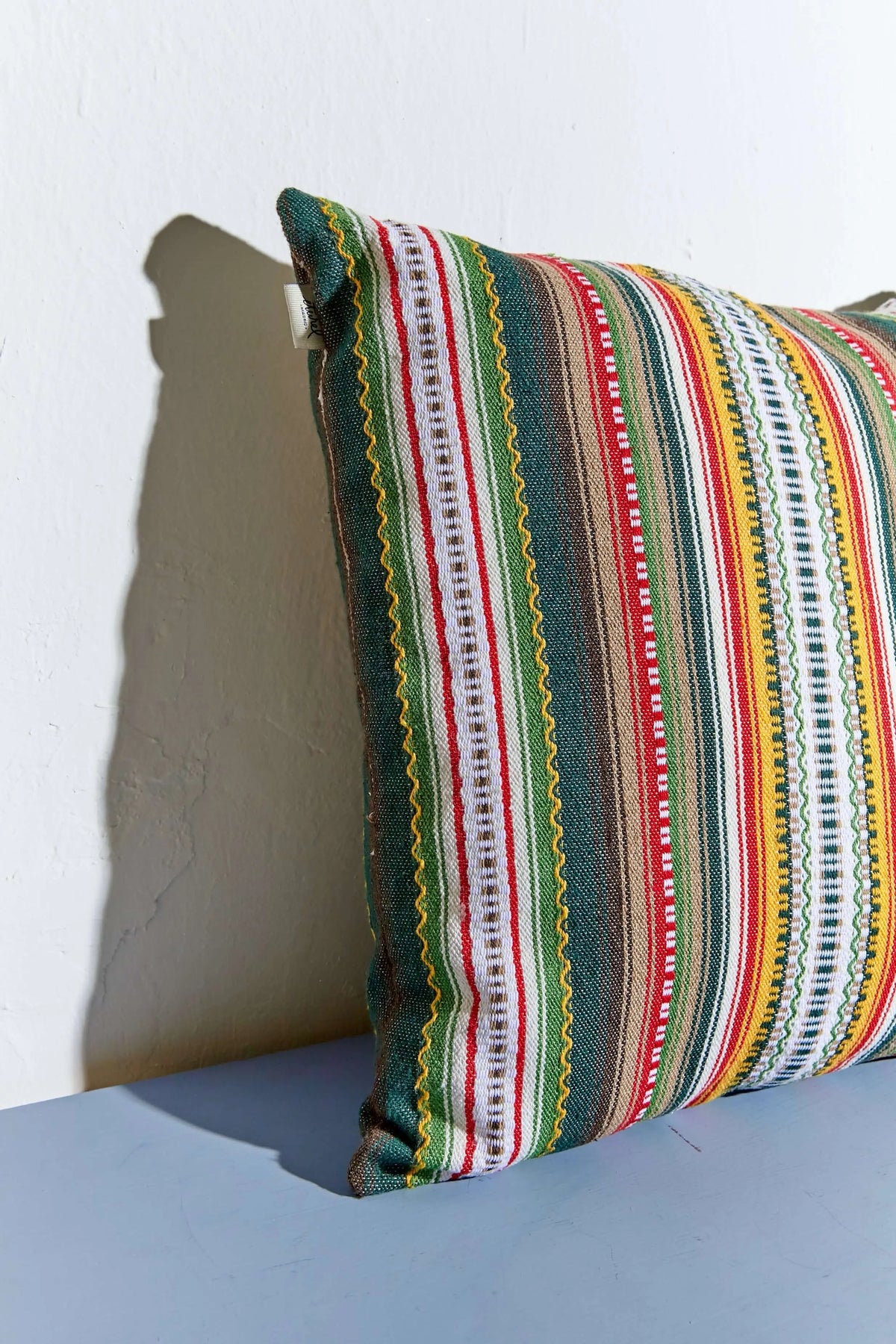 Cojín cuadrado de tela tejida con rayas verticales en tonos verde, rojo, amarillo y blanco sobre fondo neutro.